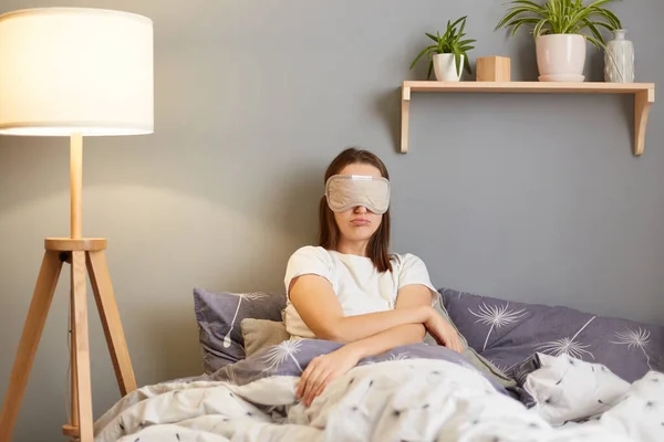 Indoor shot of attractive sad upset woman wearing pajama and sleeping mask, sitting in bed, expressing sorrow and sadness, needs to have nap, doesn' t want to get up.