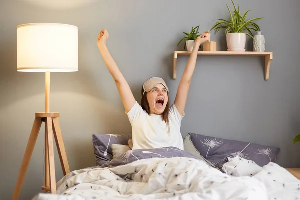 Indoor shot of young adult satisfied Caucasian woman with sleeping mask lying in bed, waking up after good sleep, stretching her arms with happy expression.