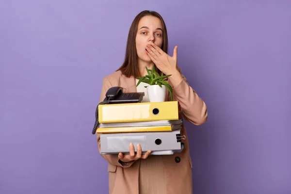 Portrait of shocked surprised woman wearing beige jacket holding lot of documents folders isolated over purple background, looking at camera, covering mouth with palm, having mistake at her work.