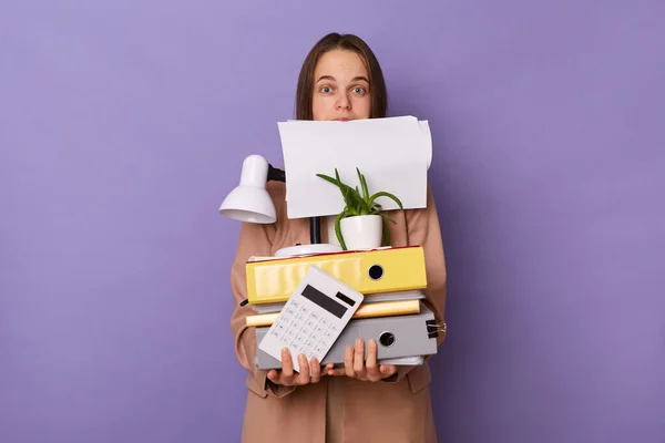 Portrait of busy funny woman with papers in mouth wearing beige jacket holding lot of documents folders isolated over purple background, looking at camera with big eyes.