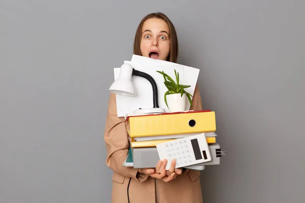 Indoor shot of shocked astonished woman wearing beige jacket posing isolated over gray background, having lots of work, looking at camera with big eyes, holding paper folders.