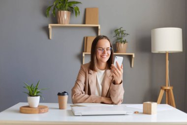Kapalı alanda, kahverengi saçlı, bej ceketli, olumlu iyimser bir kadının ofiste çalışırken, kocasıyla ya da arkadaşıyla görüntülü konuşma yaparken görüntüsü var..