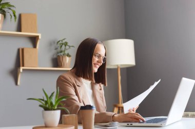 Kahverengi saçlı, bej ceketli güzel bir kadının yan görünüm portresi. İnternette çalışıyor, iş yerinde oturuyor ve elinde kağıt belgeler var..