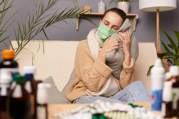 Indoor shot of sleepy tired Caucasian young adult woman wearing warm sweater, scarf and protective mask sitting on sofa in home interior among pills, holding pillow in hands and having nap.