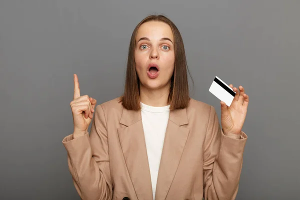Indoor shot of excited surprised woman with brown hair wearing beige jacket posing isolated over gray background, keeps mouth widely open, raised finger up, having idea how to spend money on card.