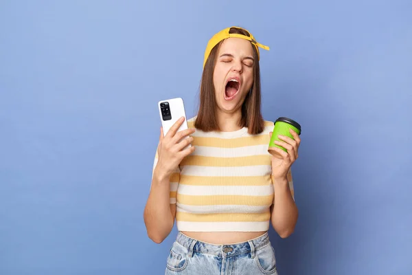 Horizontal shot of sleepy tired teen girl wearing striped T-shirt and baseball cap standing isolated over blue background, holding coffee to go and cell phone, yawning, needs energy.