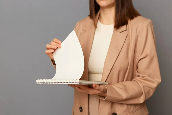Portrait of anonymous faceless woman wearing beige official style jacket standing isolated over gray background, holding paper documents, turning pages, working.