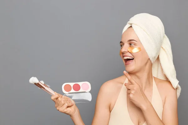 Horizontal shot of satisfied delighted woman wearing towel and with patches under eyes, holding eye shadow palette and brushed and pointing away at copy space for advertisement.