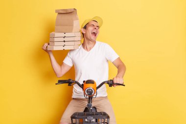 Indoor shot of crazy silly overjoyed delivery man with pizza in cardboard box, sitting on fast motorbike isolated over yellow background, laughing out loud.