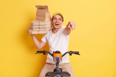 Image of courier man delivers tasty fresh baked pizza, wishes good appetite for you, holds cardboard box, wears white t-shirt, sits on fast motorbike, pointing to camera isolated on yellow wall.