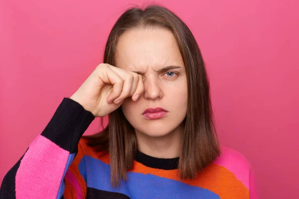 Horizontal shot of sad tired stressed woman wearing colorful jumper posing isolated over pink background, looking at camera and crying, rubbing her eye, feels pain.
