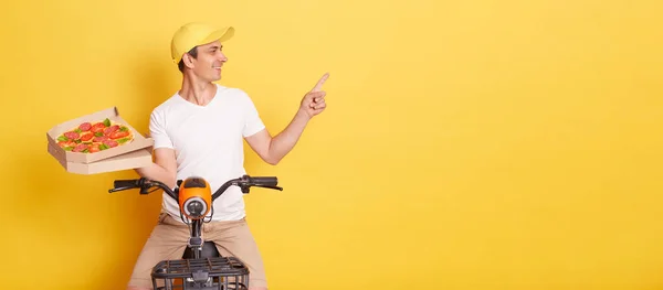 Photo of delivery man with pizza in cardboard box, wearing white t-shirt, sitting on fast motorbike and pointing at copy space for advertisement, posing isolated over yellow background.