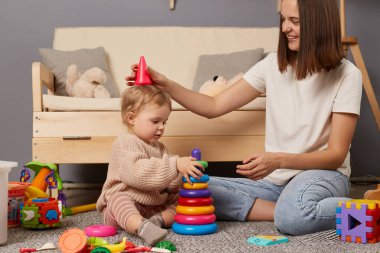 Image of dark haired mother and daughter playing with pyramid, having fin together while sitting on floor, early childhood development, educational games.