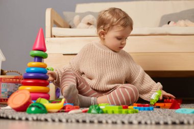 Horizontal shot of baby wearing stylish sweater playing with colorful rainbow toys, sitting on floor with attentive look, educational toys for children, early development.