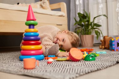 Portrait of smiling charming adorable lying on floor, being tired to play, posing near sofa and colorful pyramid, looking at camera, being surrounded different toys.