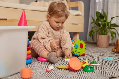 Indoor shot of concentrated baby playing with colorful rainbow sorter toy, sitting on floor near sofa, toys for little kids, interior for little girl nursery.