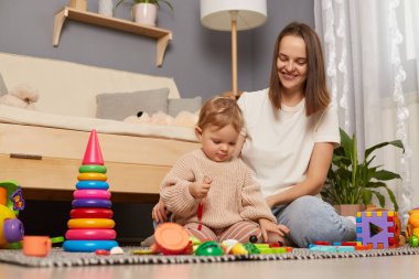 Indoor shot of Caucasian woman wearing white t-shirt sitting on floor with her charming daughter, playing with colorful toys, early development, expressing happiness.