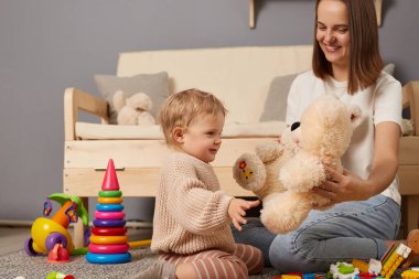 Indoor shot of smiling positive optimistic cheerful cute baby and mother sitting on floor at home and playing with plush teddy bear, smiling happily, enjoying time spend together.