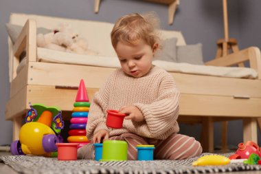 Image of cute charming little baby girl playing with colorful toys at home sitting on floor, playing early education games, learning colors and sizes, having concentrated expression.