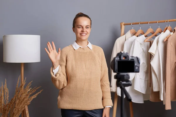 Horizontal shot of smiling beautiful Caucasian wearing beige jumper recording video in her showroon for her blog about fashion, waving hand, saying hello to followers.