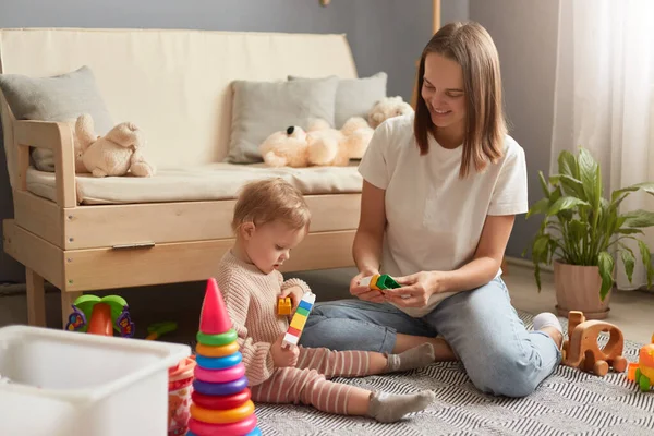 Indoor shot of mother and baby daughter playing with colorful constructor blocks while sitting on the floor in living room at home, family spending time together.