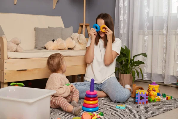 Activities with children at home, early childhood development. Young adult woman sitting on floor in living room, playing with her baby child with pyramid, covering her eyes with rings, having fun.