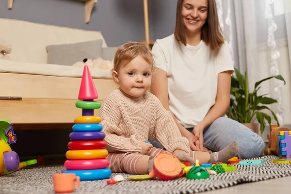 Indoor shot of mother and toddler daughter playing on the floor early childhood development, family spending time together with colorful pyramid, expressing happiness.