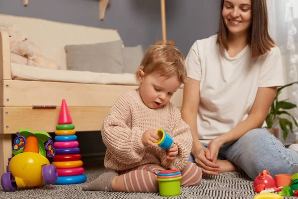 Indoor shot of beautiful mom plays with baby in the room with colorful plastic toys, happy time together, early development, mommy enjoying her maternity leave.