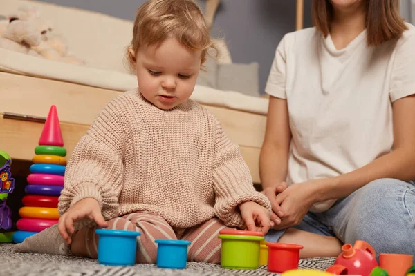Early development. Photo of little toddler girl in beige sweater playing with educational colorful toys at home, sitting in living room near sofa with her mother.
