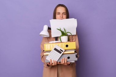 Portrait of busy funny Caucasian woman wearing official style jacket holding folders isolated over purple background, holds empty papers with her teeth, looking at camera.