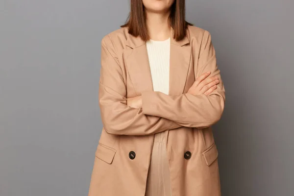 Fashionable women\'s outfit. Woman posing in white top and beige jacket and trousers, faceless female with brown hair isolated over gray background, keeps hands folded, business style.