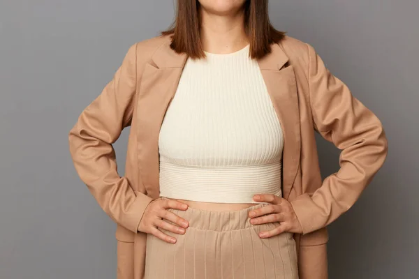 Fashionable women's outfit. Woman posing in white top and beige jacket and trousers, faceless female with brown hair isolated over gray background, keeps hands in on hips, business style.