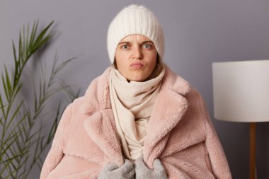 Horizontal shot of sad serious woman wearing cap and winter coat, looking at camera with pout lips and disappointed facial expression,having problems with heating at home.