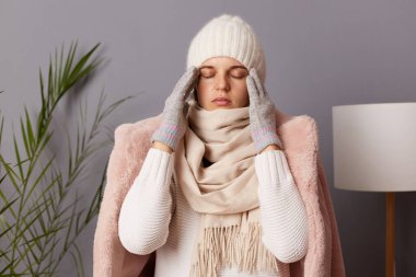 Indoor shot of sick unhealthy woman wearing cap and winter coat sitting on the sofa at home, having flu or grippe symptoms, suffering terrible headache, catching cold in cold house.