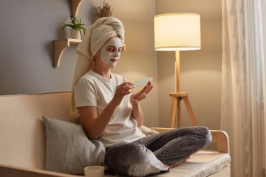 Indoor shot of attractive relaxed woman with cosmetology mask on face sitting on cough, doing anti aging procedures and manicure, posing in lotus pose, beauty routine.