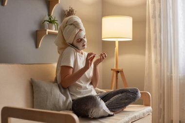 Photo of attractive woman wearing white T-shirt and pajama pants, wrapped in white towel sitting on sofa with cosmetic mask, doing manicure and talking with friends.