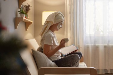 Horizontal shot of woman wearing white T-shirt and in towel sitting on cough near lamp and reading book, holding cup of coffee, enjoying time on weekend, applying cream for rejuvenation.