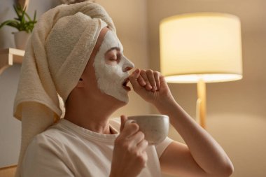Profile portrait of woman in white towel sitting on sofa at home doing cosmetic procedures, holding coffee cup, feels sleepy, yawning, covering mouth with hand.