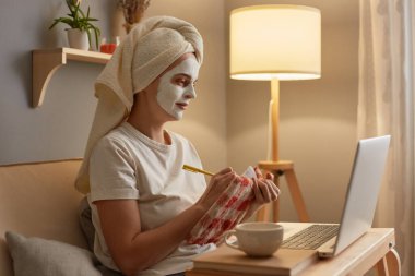 Portrait of Caucasian woman in white towel sitting on sofa at home doing cosmetic procedures, writing in notebook working online on laptop, female freelancer works at home.
