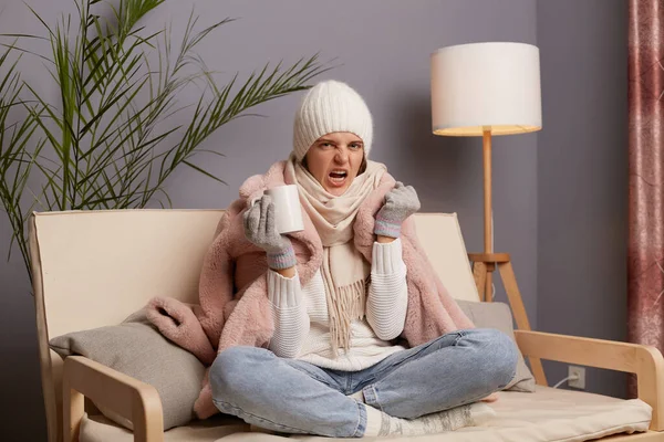 Indoor shot of angry aggressive woman wearing cap and winter coat sitting on the sofa at home, trying to get warm in cold apartment, holding cup with hot tea, clenched fist.
