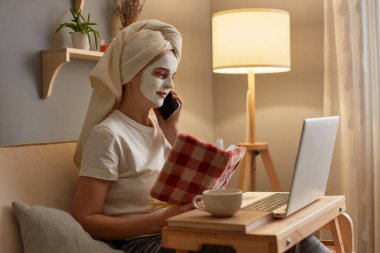 Portrait of adorable woman wearing white T-shirt and in towel sitting on cough near lamp, doing skin care procedures, holding organizer, looking at laptop display and talking with boss on cell phone.
