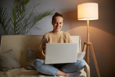 Portrait of delighted attractive woman freelancer wearing beige jumper and jeans sitting on cough and working online at home, holding laptop, typing on keyboard.