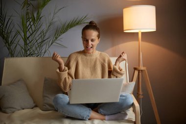 Indoor shot of joyful cheerful excited amazed woman wearing beige jumper and jeans, clenched fists while looking at laptop display, sitting on cough with crossed legs.