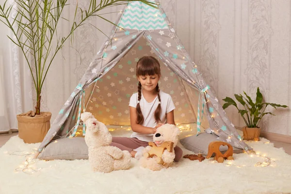 Indoor shot of adorable little girl with pigtails wearing white t-shirt playing with toys in teepee tent, holding soft rabbit, playing games, trying to have fun alone.