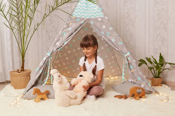 Indoor shot of charming joyful little girl with pigtails wearing white t-shirt playing with toys in wigwam, holding soft teddy bear, playing alone with her favorite toy.