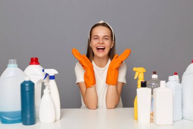 Horizontal shot of joyful cheerful woman wearing protective gloves, sits at table with many detergents, posing isolated over gray background, screaming with happiness, enjoying household chores.