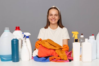Caucasian smiling satisfied woman wears rubber protective gloves and headband, surrounded with bottles of detergents for laundry, isolated over gray background, looking at camera.