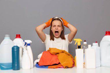 Image of shocked yelling sad woman wears headband and casual t shirt, sits at workplace with different detergents for laundry and lots dirty clothes, isolated over gray background, screaming.