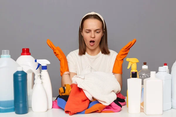 Indoor shot of shocked sad housewife, wears rubber protective gloves, surrounded with stain remover, spray and other chemical products for laundry, has lots of work, being upset.