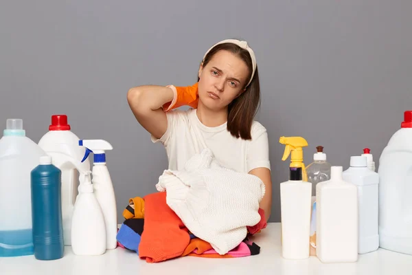 Horizontal shot of tired exhausted woman wearing gloves, sits at table with many detergents and dirty linen, needs doing laundry, feels fatigue and pain neck, posing isolated over gray background.